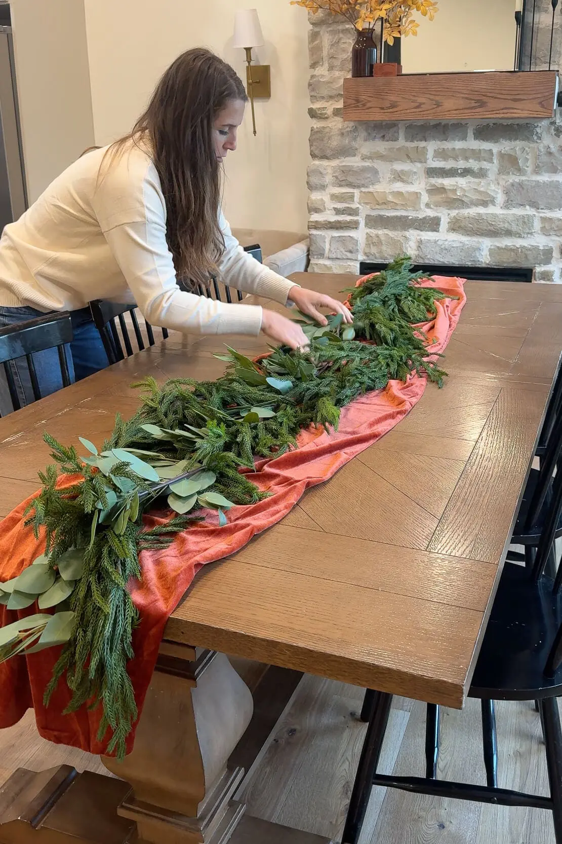 Woman arranging fresh greenery, including pine branches and eucalyptus, along an orange velvet table runner for a holiday table setting.